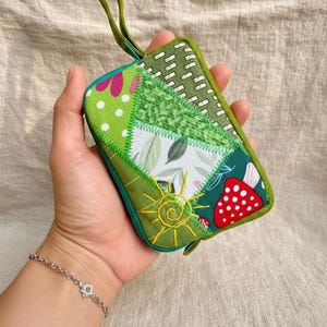 May include: A handmade patchwork wallet in shades of green and white, featuring floral and mushroom patterns, and a yellow embroidered sun. It has a green strap. The wallet is approximately 15 cm long.