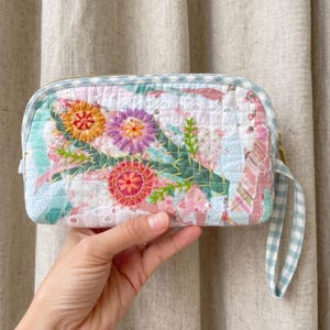 May include: A quilted pouch with a floral embroidery design. The pouch features a mix of pastel colors, including pink, blue, and green, with a gold zipper and a blue and white gingham wrist strap. The embroidery includes orange, purple, and red flowers.