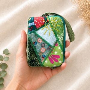 May include: A small rectangular zippered pouch featuring a patchwork design of various green, blue, and red fabrics. The front panel displays embroidered details including a yellow sun, floral patterns, and polka dots, finished with a green wrist strap.