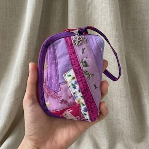 May include: A small, patchwork wallet in shades of purple, pink, and white. The wallet has a zipper closure and a small loop handle. A silver clover charm hangs from the zipper pull. The fabric features floral and geometric patterns.