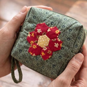 Op de afbeelding: Een vierkant, gewatteerd etui met een bloemmotief. Het etui is groen met een herhalend bloemmotief en een grote, geappliqueerde bloem in rood, geel en wit. Het heeft een ritssluiting en een kleine stoffen lus.