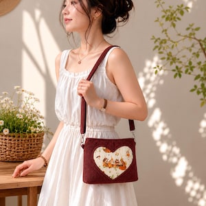 May include: A burgundy corduroy crossbody bag with a heart-shaped patch featuring a whimsical illustration. The bag has a matching burgundy strap and silver-tone hardware. The background is a soft, neutral color.