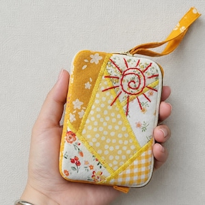 Könnte beinhalten: Eine rechteckige Stofftasche mit Reißverschluss und Patchwork-Design in Gelb- und Weißtönen. Die Außenseite zeigt Blumen-, Punkte- und Vichy-Karomuster mit einem handgestickten roten Sonnenmotiv. Eine gelbe Stoffschlaufe ist angebracht.
