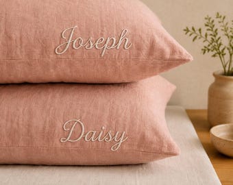 Könnte beinhalten: Zwei rechteckige Kissen aus altrosa Leinenstoff, die übereinander gestapelt sind. Das obere Kissen ist mit dem Namen Joseph in weißer Schreibschrift bestickt, das untere Kissen mit dem Namen Daisy in weißer Schreibschrift.