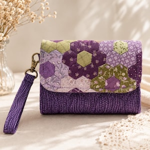 May include: A quilted fabric wristlet featuring a flap with a hexagonal patchwork pattern in shades of purple, lavender, and olive green. The main body is a deep purple fabric with a subtle wavy texture, and it includes a matching wrist strap.