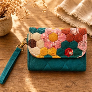 May include: A teal quilted wallet with a colorful patchwork flap featuring a floral and geometric design. The wallet has a teal wrist strap and a gold-colored clasp. The patchwork includes shades of pink, yellow, brown, and green.