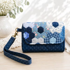Puede incluir: Un bolso de mano de tela acolchada con una solapa de patrón de patchwork hexagonal azul y blanco con estampados florales y geométricos. El cuerpo principal es azul oscuro con un patrón de círculos blancos y una correa de muñeca.