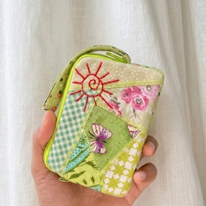 May include: A patchwork wallet with a variety of fabric patterns, including floral, gingham, and geometric designs. The wallet features a hand-stitched sun and butterfly applique. The color palette includes shades of green, pink, and white. It has a wrist strap and a yellow zipper.
