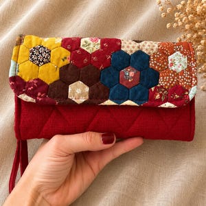 Può includere: Un portafoglio rosso trapuntato con patta patchwork colorata. Il patchwork presenta pezzi di tessuto esagonali in giallo, rosso, marrone e blu, creando un motivo floreale. Il portafoglio ha un cinturino da polso.