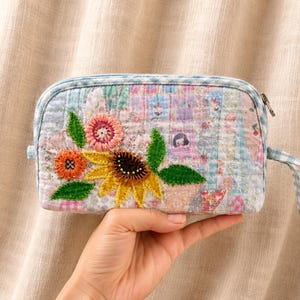 Könnte beinhalten: Kleine, gesteppte Tasche mit floraler Stickerei. Die Tasche hat ein Patchworkmuster in Pastellfarben und eine blau-weiß karierte Bordüre. Die gestickten Blumen umfassen eine Sonnenblume, eine orangefarbene Blume und eine rosa Blume mit grünen Blättern. Ein kleiner Reißverschluss und eine Handschlaufe sind ebenfalls sichtbar.