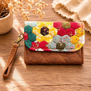 Può includere: Un portafoglio trapuntato marrone con patta patchwork colorata. La patta presenta un motivo di pezzi di tessuto esagonali in vari colori, tra cui giallo, rosso, verde e stampe floreali. È attaccato un cinturino da polso marrone con chiusura color oro.