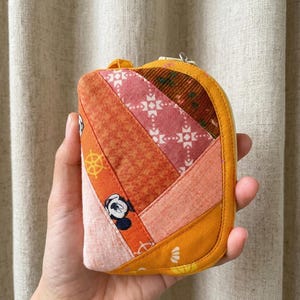 May include: A small, colorful, patchwork zippered pouch. The pouch features a variety of fabrics in shades of orange, pink, and yellow, with a Mickey Mouse print and geometric patterns. The pouch is held in a hand.