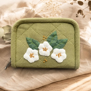May include: A quilted, olive green wallet with a zipper closure. The front features three white flower appliqués with gold bead centers and green leaf accents. The wallet is approximately 5 inches wide.