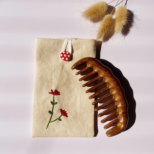 May include: A natural-coloured canvas pouch featuring a red and white polka dot button closure and an embroidered red flower design, accompanied by a wide-toothed wooden comb with a visible grain pattern and three dried beige grass plumes.