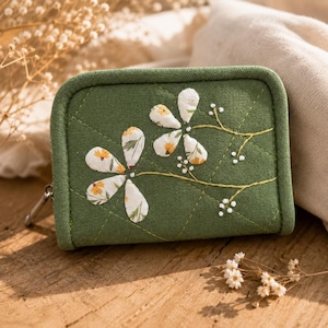 May include: A green quilted wallet with a floral design. The wallet features two large white flowers with yellow and green accents, and delicate embroidered stems and white dots. The wallet has a zipper closure and a rounded shape.