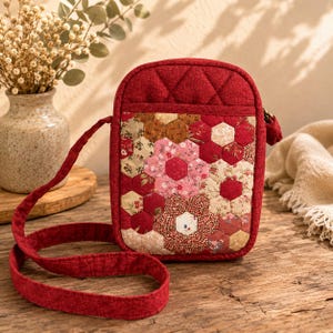 May include: A red quilted crossbody bag with a floral patchwork design. The bag has a long, matching strap and a zip closure. The patchwork incorporates various floral patterns in shades of red, pink, and beige. The bag is set on a wooden surface.