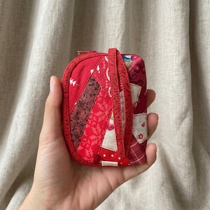 Könnte beinhalten: Eine kleine, rechteckige Stofftasche mit einem Patchwork-Design in verschiedenen Rottönen. Die Außenseite besteht aus mehreren gesteppten Stoffstreifen mit Blumen- und geometrischen Mustern, inklusive einer passenden roten Handschlaufe und Reißverschluss.