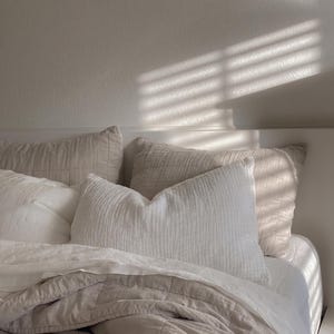 K&ouml;nnte beinhalten: Ein Bett in einer Farbpalette aus Wei&szlig; und hellem Beige mit mehreren Kissen und einer gesteppten Decke. Die Bettw&auml;sche umfasst ein strukturiertes wei&szlig;es Kissen, zwei beige gesteppte Kissen und eine beige gesteppte Decke &uuml;ber wei&szlig;en Laken.