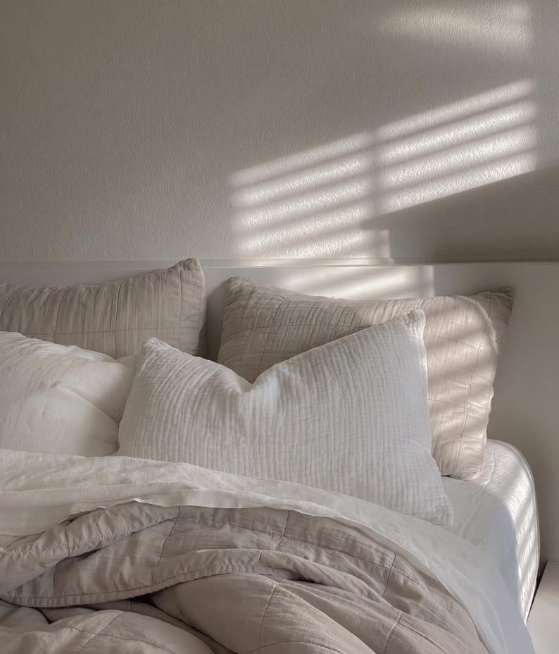 K&ouml;nnte beinhalten: Ein Bett in einer Farbpalette aus Wei&szlig; und hellem Beige mit mehreren Kissen und einer gesteppten Decke. Die Bettw&auml;sche umfasst ein strukturiertes wei&szlig;es Kissen, zwei beige gesteppte Kissen und eine beige gesteppte Decke &uuml;ber wei&szlig;en Laken.