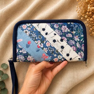 Può includere: Un portafoglio trapuntato con finiture e cinturino da polso blu navy. Il portafoglio presenta un design patchwork con motivi floreali e geometrici nei toni del blu, bianco e rosa. La cerniera è argentata.