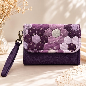May include: A purple quilted wallet featuring a hexagonal patchwork pattern in various shades of violet and lavender. The wallet includes a matching purple fabric wrist strap attached with a bronze-toned metal lobster clasp and a fold-over flap closure.