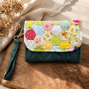 Könnte beinhalten: Eine handgefertigte Geldbörse mit Patchwork-Design. Der Umschlag hat bunte sechseckige Stoffstücke mit floralen und geometrischen Mustern. Der Körper ist aus dunkelgrünem, gestepptem Stoff. Inklusive grüner Handschlaufe und bronzefarbenem Verschluss.