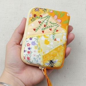 Könnte beinhalten: Ein Patchwork-Etui mit Reißverschluss und Bienen- und Blumenmuster. Das Etui hat gelbe, orangefarbene und weiße Stoffe mit Blumen- und Beerenmustern. Ein kleiner orangefarbener Stoffzug ist am Reißverschluss befestigt.