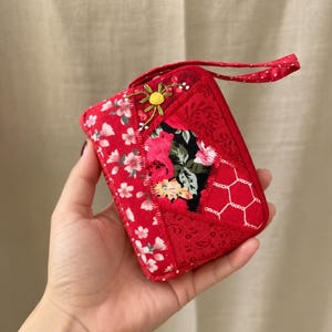 Puede incluir: Un pequeño estuche rojo con cremallera que presenta un diseño de patchwork con varios patrones de tela florales y geométricos. El estuche incluye una correa de muñeca roja a juego con lunares blancos y un pequeño detalle de flor bordada.