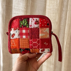Puede incluir: Un pequeño monedero cuadrado acolchado con un diseño de patchwork. El monedero presenta una variedad de telas rojas con estampados de cerezas y flores. Tiene una cremallera roja y una pequeña correa de muñeca. El monedero se sostiene en una mano.
