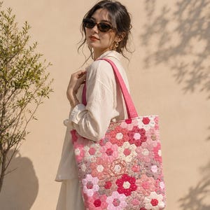 Peut inclure: Un sac fourre-tout matelassé présentant un motif patchwork composé de pièces de tissu hexagonales dans diverses nuances de rose, rouge et blanc à motifs floraux. Le sac possède deux anses roses unies et une forme rectangulaire surpiquée.