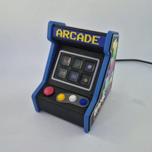 May include: A small arcade machine in blue and black, with the word "ARCADE" in yellow pixelated text. It has six buttons with app icons and four coloured buttons below. The design is reminiscent of classic arcade games.
