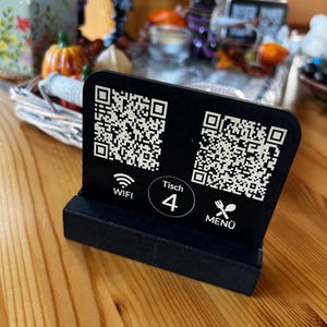 Könnte beinhalten: Ein schwarzes Tischschild mit zwei QR-Codes, einem WLAN-Symbol und dem Wort "WIFI". Das Schild zeigt auch die Nummer "4" in einem Kreis und das Wort "MENU" mit einem Gabel- und Messersymbol. Das Schild steht auf einem Holztisch.