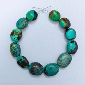 Könnte beinhalten: Eine Kette aus türkisfarbenen Edelsteinperlen. Die oval geformten Perlen weisen eine Mischung aus Türkis, Braun und Schwarz auf. Sie sind mit einem dünnen, silberfarbenen Draht aufgefädelt. Die Perlen sind kreisförmig angeordnet.