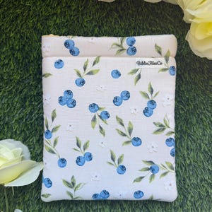 May include: A rectangular fabric sleeve featuring a repeating pattern of blue blueberries, green leaves, and small white flowers on an off-white background. A small white fabric tag with the black text BiblioFilesCo is sewn onto the upper right side.