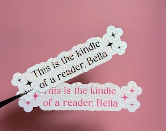 May include: Two holographic stickers with the text "This is the kindle of a reader, Bella" in black and pink. The stickers have a sparkly, star-like design around the edges and are set against a pink background.