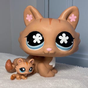 Peut inclure: Une grande figurine de chat marron avec des rayures orange, de grands yeux noirs avec des pupilles en forme de fleur blanche et des motifs de fleurs roses sur les oreilles, accompagnée d'une figurine de chat plus petite assortie.