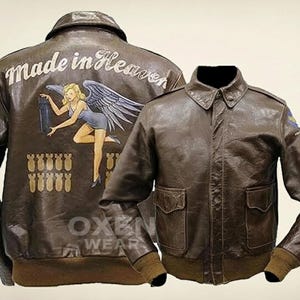 Könnte beinhalten: Braune Leder-Bomberjacke mit klassischem Kragen und zwei Vordertaschen. Auf der Rückseite der Jacke befindet sich der Text "Made in Heaven" über einem gemalten Bild einer Frau mit Flügeln, die eine Bombe hält. Die Jacke hat einen gerippten Saum und Bündchen.