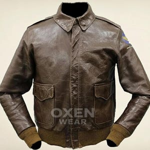 Op de afbeelding: Bruine leren bomberjack met een kraag, twee voorzakken en geribde manchetten en zoom. De jas heeft een patch op de bovenarm en de tekst "OXEN WEAR" op de voorkant.