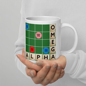 May include: White ceramic mug featuring a Scrabble-themed design. The mug displays a green game board with the word "OMEGA" and "ALPHA" spelled out using Scrabble tiles. Includes bonus score spaces.