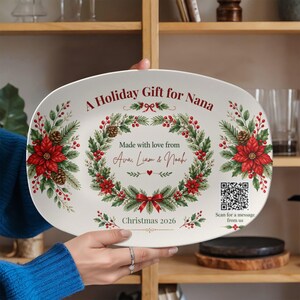 May include: An oval white ceramic serving platter featuring a festive wreath design with red poinsettias, pine needles, and berries. The text reads A Holiday Gift for Nana, Made with love from Ava, Liam & Noah, Christmas 2026, and includes a QR code.