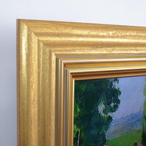 Op de afbeelding: Een ingelijst landschapsschilderij met een goudkleurige, sierlijke lijst. Het schilderij toont een groen veld, bomen en een blauwe lucht. De lijst heeft een textuuroppervlak en een gelaagd ontwerp.