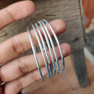 May include: A set of six thin, silver-colored bangles. The bangles are held in a hand, with a wooden surface in the background. The ends of the bangles have a blue hue. The text "925SilverArtsCo" is visible in the upper right corner.