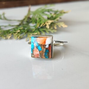 May include: A square-faced ring with a mosaic of turquoise, orange, white, and brown stones set in a silver band. The ring is displayed on a white surface with a blurred green plant in the background. The text "925SilverArtsCo" is visible.