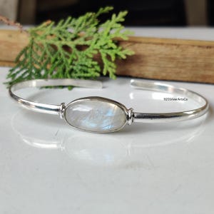 Pode incluir: Uma pulseira de punho de prata com um centro de pedra da lua oval e iridescente. A pulseira é aberta e apresenta o texto "925SilverArtsCo". A pedra da lua exibe um jogo de cores, refletindo a luz. A pulseira é provavelmente feita de prata esterlina.