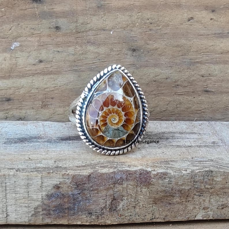 K&ouml;nnte beinhalten: Ein tropfenf&ouml;rmiger Ring mit einem polierten Ammoniten-Fossil mit braunen und beigen Spiralmustern. Der Stein ist in ein silbernes Band mit einer gedrehten Kordelbord&uuml;re gefasst. Der Text 925SilverLegance ist auf der rechten Seite des Rings sichtbar.