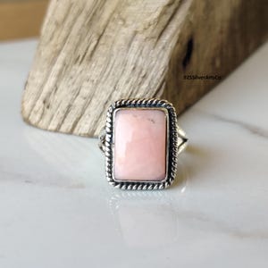 May include: A silver ring with a rectangular, light pink gemstone. The gemstone is set in a silver frame with a twisted rope design. The ring is displayed on a white surface.