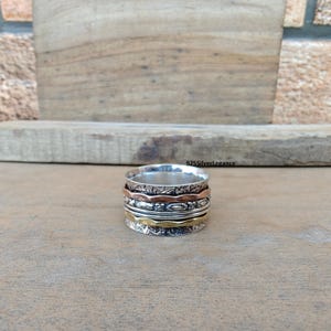 May include: A silver spinner ring with copper and gold-colored bands. The ring has a detailed floral pattern on the top and bottom bands. The ring is displayed on a wooden surface.