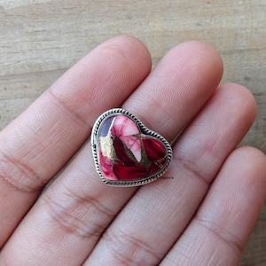 May include: A heart-shaped ring with a silver band. The heart is a mix of red, pink, and gold-colored stone. The ring is held in a hand, and the background is a light brown wooden surface.