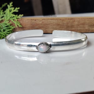 May include: A silver cuff bracelet featuring an oval opal gemstone set in a textured bezel. The bracelet has a smooth, polished finish and an open design. The jewellery is displayed on a white surface.