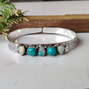 Puede incluir: Un brazalete de plata con una superficie texturizada. El brazalete está adornado con cinco piedras preciosas: dos ópalos ovalados y tres piedras turquesas. El brazalete está abierto en la parte superior y se muestra sobre una superficie blanca.